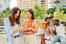 Two attractive and cheerful smiling women looking and showing the last funny and entertaining news on their smartphone outside at the university campus. At background, three young friends, having fun taking selfies in free time. Lifestyle concept